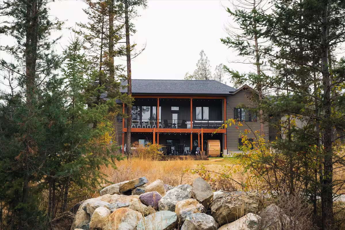 Back of the home framed by pines with a two-level covered deck and barrel sauna