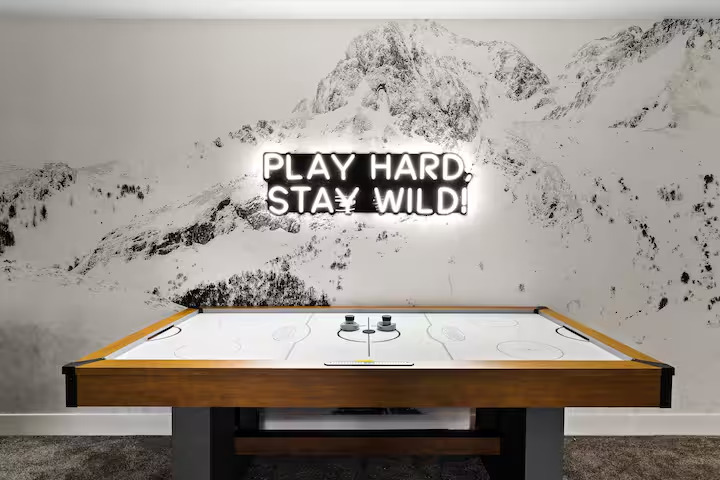 Air hockey table beneath a black-and-white mountain mural and a Play Hard Stay Wild neon sign