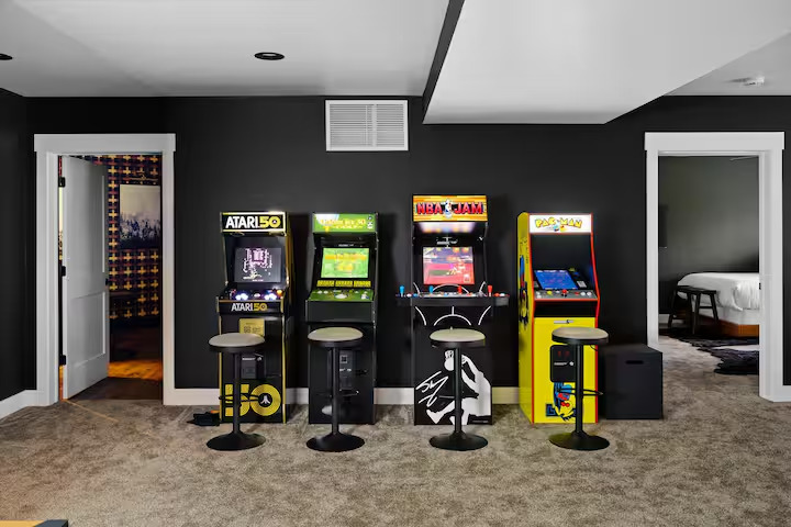 Row of retro arcade cabinets with bar stools along a charcoal wall in the game room